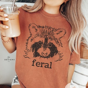 Puede incluir: Camiseta de color terracota con un gráfico negro de un mapache y la palabra "feral" impresa debajo. La camiseta tiene un corte holgado y mangas cortas. La imagen también muestra a una persona sosteniendo un vaso de café helado.