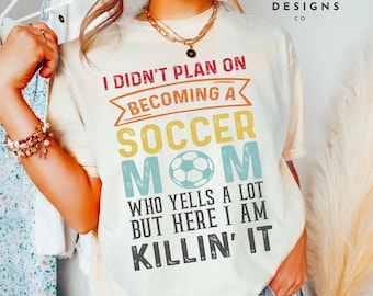 May include: A white t-shirt with a vintage-style graphic that reads "I didn't plan on becoming a soccer mom who yells a lot but here I am killin' it".