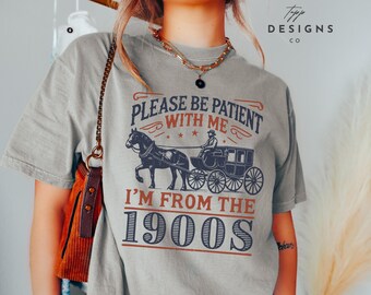 Puede incluir: Una camiseta gris con un gráfico de estilo vintage que dice "Por favor, ten paciencia conmigo, soy de los años 1900". El gráfico presenta un carruaje tirado por caballos con un conductor.