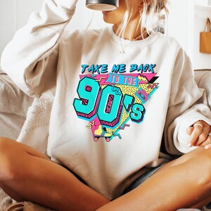 Puede incluir: Una sudadera blanca con un estampado gr&aacute;fico que dice "Take Me Back To The 90's" en letras coloridas de estilo retro. El gr&aacute;fico tambi&eacute;n incluye un pat&iacute;n y otros dise&ntilde;os inspirados en los 90.