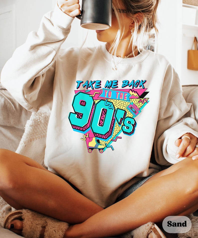 Puede incluir: Una sudadera blanca con un estampado gr&aacute;fico que dice "Take Me Back To The 90's" en letras coloridas de estilo retro. El gr&aacute;fico tambi&eacute;n incluye un pat&iacute;n y otros dise&ntilde;os inspirados en los 90.