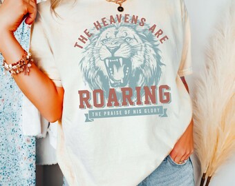 May include: A light beige t-shirt with a vintage-style graphic of a roaring lion. The text reads "The Heavens Are Roaring, The Praise Of His Glory".