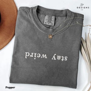 Puede incluir: Una camiseta gris oscuro con el texto "Stay Weird" impreso en blanco en la parte delantera. La camiseta está hecha de un material suave y cómodo y tiene un corte holgado.