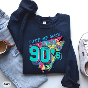 Puede incluir: Una sudadera azul marino con un estampado gr&aacute;fico del texto "Take Me Back To The 90's" en un estilo retro colorido. El gr&aacute;fico incluye una cinta de casete, patines y formas geom&eacute;tricas.