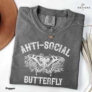 Puede incluir: Una camiseta gris oscura con un gráfico de mariposa blanca y el texto "Anti-Social Butterfly".