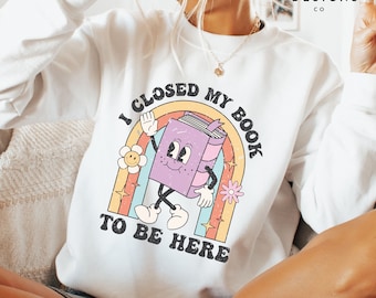May include: White sweatshirt with a rainbow and a cartoon book character. The text on the sweatshirt reads "I closed my book to be here."