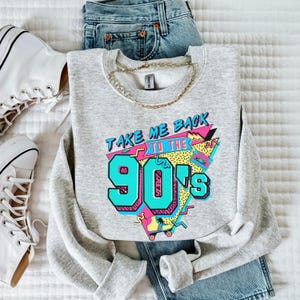 Puede incluir: Una sudadera gris con un estampado gr&aacute;fico que dice "Take Me Back To The 90's" en un estilo retro colorido. El gr&aacute;fico incluye una cinta de casete, patines y formas geom&eacute;tricas.