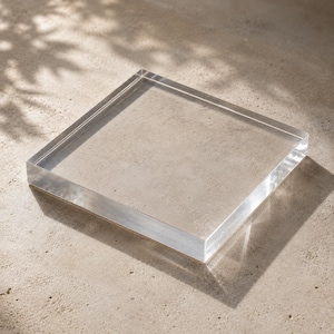 May include: A square, clear acrylic block with smooth, polished edges and a flat top surface. The transparent material allows for a clear view through the object, which features sharp, defined corners and a solid, uniform construction throughout.