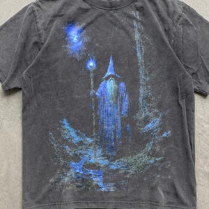 May include: A charcoal gray short-sleeve t-shirt featuring a graphic print of a robed figure in a pointed hat holding a glowing blue staff. The figure stands in a dark, wooded landscape with blue light accents and a bright blue star above.