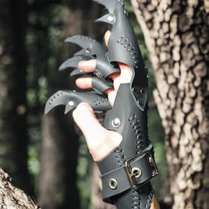 May include: Black leather claw gloves with curved, pointed tips and stitched detailing. The gloves have a wrist strap with a buckle and eyelets. The background is a blurred view of a forest.