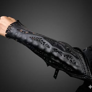 Puede incluir: Guardabrazos de cuero negro con cortes decorativos y ojales metálicos. El guardabrazos está diseñado para proteger el antebrazo y la muñeca. El cuero es texturizado y tiene un acabado mate. Incluye una correa y hebilla para una sujeción segura.