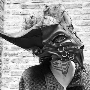 May include: A black leather mask with intricate details, including stitching, rivets and metal rings. The mask has a long, pointed snout and large, pointed ears. The mask is worn by a person with curly hair.