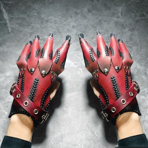 Può includere: Un paio di guanti in pelle rossa con dita lunghe e appuntite. I guanti presentano cuciture nere, borchie metalliche e cinturini da polso con fibbie. Il design ha un tema gotico o fantasy.