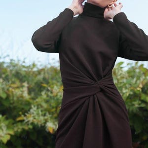 Puede incluir: Vestido marrón oscuro de manga larga con cuello alto y detalle de nudo en la cintura. El vestido está hecho de una tela suave y fluida. La modelo lleva pendientes dorados.