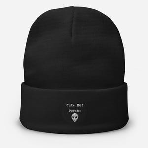 May include: Black beanie hat featuring the embroidered phrase "Cute But Psycho" and an alien head design. The hat is a plain black colour with a folded brim. A casual accessory.