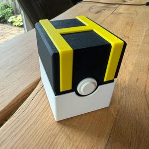 Op de afbeelding: Een kubusvormige opbergdoos met een zwart bovengedeelte met een felgele H-vormige accent, een wit ondergedeelte en een ronde witte knop in het midden van de voorzijde. De doos heeft een gestructureerde afwerking en een geometrisch ontwerp.