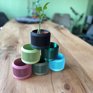 Op de afbeelding: Een stapel van zes cilindrische plantenpotten met verticale lamellen, met een matte afwerking in zwart, lichtgroen, donkergroen, roze, blauwgroen en blauw. Een kleine groene plant in een bruine kweekpot staat in de bovenste zwarte pot.