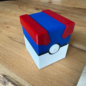 Op de afbeelding: Een vierkante opbergdoos ontworpen om op een Great Ball uit de Pokémon-serie te lijken. De container heeft een witte basis, een blauw middengedeelte met een zwarte horizontale band en een rood deksel. Een witte knop is aan de voorzijde.