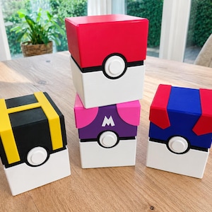 Op de afbeelding: Vier vierkante opbergdozen ontworpen als Pokeballs. Eén is rood en wit, één is zwart en geel met een H, één is paars en roze met een M, en één is blauw en rood. Elke doos heeft een centrale witte ronde knop op een zwarte band.