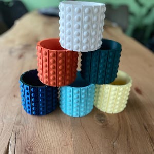 Può includere: Una collezione di sei vasi cilindrici disposti a piramide. Ogni vaso presenta un esterno strutturato con file verticali di protuberanze circolari in rilievo. I colori includono bianco, arancione, verde scuro, blu scuro, azzurro e giallo pallido.