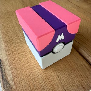 Op de afbeelding: Een vierkante opbergdoos met een paars en roze deksel met een witte letter M aan de voorzijde. De basis van de doos is wit en een ronde witte knop is gecentreerd op het voorpaneel. Het ontwerp heeft een matte afwerking met zichtbare 3D-printlagen.