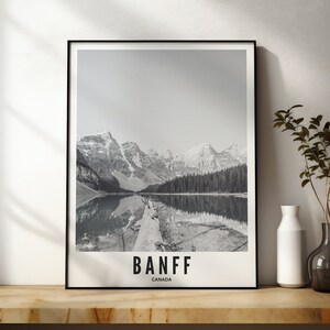 May include: Black and white framed print featuring a scenic view of Banff National Park in Canada. The image shows a mountain range reflected in a lake, with the word "BANFF" and "CANADA" printed at the bottom. The print is displayed on a wooden shelf.