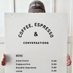 May include: A white rectangular menu sign with black text reading COFFEE, ESPRESSO & CONVERSATIONS at the top, followed by a list of drinks including Americano, Cappuccino, Double Espresso, Latte, Macchiato, Mint Chocolate, Mocha, and White Mocha.