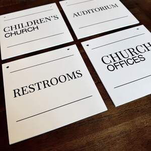 May include: Four square white signs with black text and horizontal lines. The signs are labeled Children's Church, Auditorium, Restrooms, and Church Offices. Each sign features small circular holes at the top corners for mounting purposes.