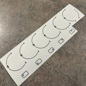 Op de afbeelding: Een set van acht witte stickers met zwarte contouren van kookplaat-branderringen. De stickers zijn ontworpen om te helpen de kookplaat te beschermen tegen krassen en slijtage.