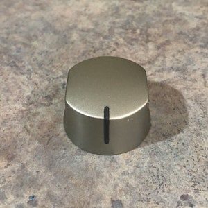 Könnte beinhalten: Ein runder, silberfarbener Herdregler mit einem schwarzen Strich in der Mitte.