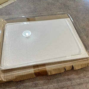 Puede incluir: Una bandeja de plástico transparente con un inserto de espuma blanca. La bandeja está sobre una caja de cartón marrón.