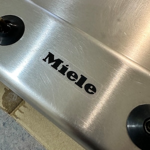 May include: Close-up of a stainless steel hob with the Miele logo in black lettering.