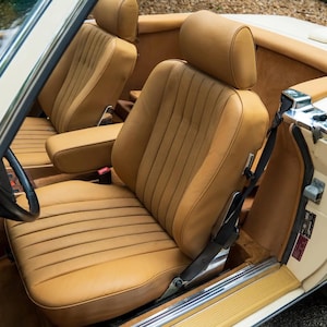May include: The interior of a convertible car featuring tan leather bucket seats with vertical stitching, a matching tan center console, a black steering wheel, and a cream-colored exterior body panel visible near the open door frame.