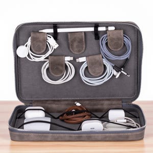 May include: Gray leather travel organizer, open to display various tech accessories. Includes a white stylus, charging cables, and charging bricks. The organizer is designed to keep electronics and accessories neatly stored.