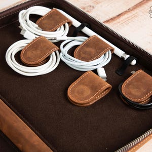 May include: A brown leather tech organizer with a zipper, containing white charging cables and a stylus. The cables are secured with small, brown leather cable organizers. The organizer has a "Home Made" stamp.