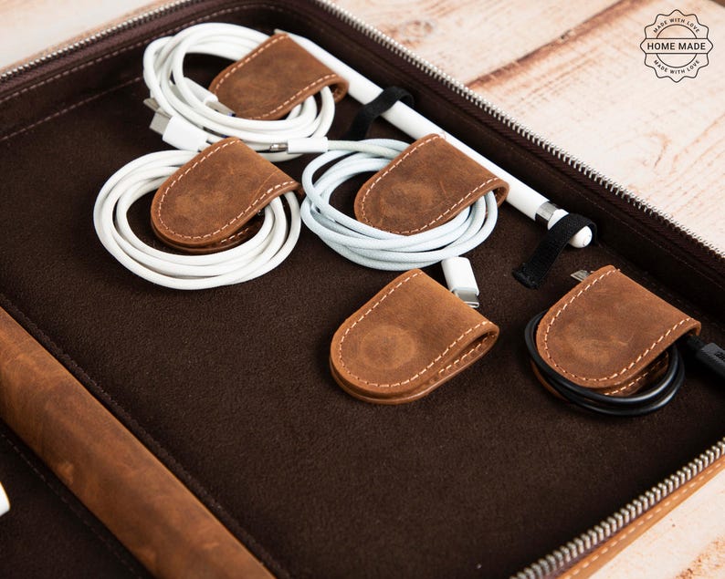 May include: A brown leather tech organizer with a zipper, containing white charging cables and a stylus. The cables are secured with small, brown leather cable organizers. The organizer has a "Home Made" stamp.