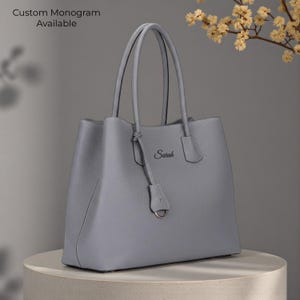 May include: A gray leather tote bag with two handles and a detachable charm. The bag has the name "Sarah" printed on the front. The text "Custom Monogram Available" is visible in the upper left corner. The bag is sitting on a round, light-colored pedestal.