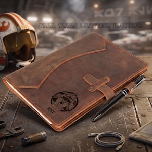May include: A brown leather journal with the text "STAR WARS LEATHER JOURNAL" and a graphic of a space station. A black pen rests on the journal. A Star Wars helmet is in the background. The journal has a strap closure and the name "SCOTT" is engraved on the cover.