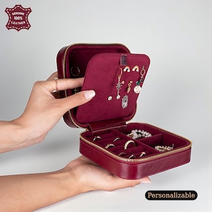 Könnte beinhalten: Eine burgunderfarbene Leder-Schmuckschatulle mit goldenem Reißverschluss, geöffnet, um Ringe, Ohrringe und Halsketten zu präsentieren. Der Innenraum ist mit einem weichen, burgunderfarbenen Stoff ausgekleidet. Die Box ist mit "Personalizable" und "Genuine 100% Leather" beschriftet.