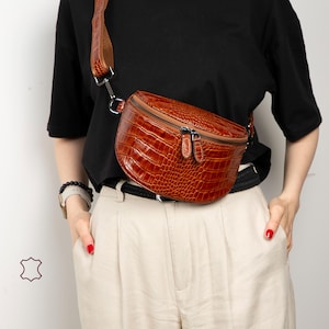 May include: A brown, textured leather fanny pack with a zipper closure and adjustable strap. The bag has a curved shape and is worn across the body. The bag is made of a crocodile-like embossed leather.