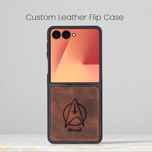 Peut inclure: Un étui à rabat en cuir personnalisé avec un fond en cuir marron orné d'un emblème de Starfleet Command et du nom "Michael" gravé en dessous. La partie supérieure affiche un écran coloré. L'étui est noir.