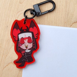 May include: A red acrylic charm depicting a cartoon character in a nun's habit with devilish features. The charm has a black clasp and a black keyring. The character has red eyes, a white habit, and a black outfit.