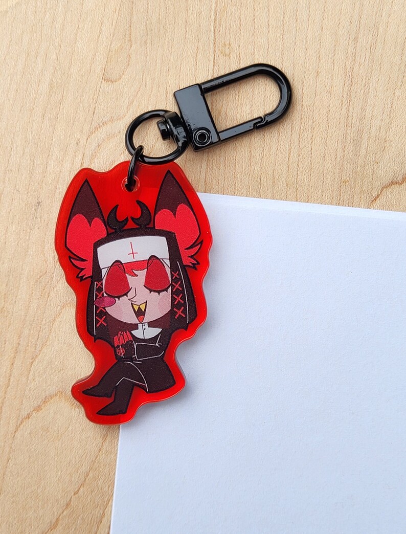 May include: A red acrylic charm depicting a cartoon character in a nun's habit with devilish features. The charm has a black clasp and a black keyring. The character has red eyes, a white habit, and a black outfit.