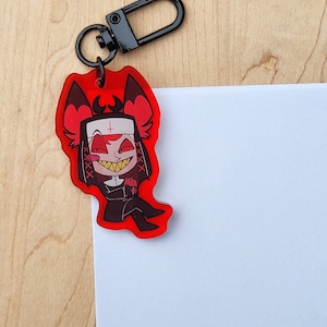 May include: A red acrylic keychain with a cartoon nun character with a devilish grin. The keychain has a black clasp. The character has red horns and a white habit with a cross. The keychain is attached to a wooden surface.