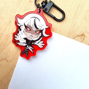 May include: A red acrylic charm with a cartoon character with white hair and red eyes. The charm has a black clasp and a black ring. The character is wearing a black outfit. The charm is on a wooden surface.