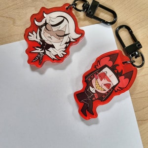 May include: Two red acrylic keyrings with black metal clasps. One keyring features a cartoon character with white hair and a black outfit. The other keyring depicts a cartoon character with red horns and a black nun's habit.