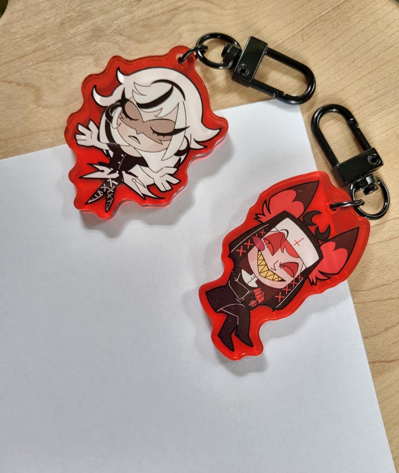 May include: Two red acrylic keyrings with black metal clasps. One keyring features a cartoon character with white hair and a black outfit. The other keyring depicts a cartoon character with red horns and a black nun's habit.