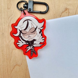 May include: A red acrylic keychain with a cartoon character design. The character has white hair and a black outfit. The keychain has a black clasp and ring. The character has closed eyes and a stern expression. The keychain is on a wooden surface.