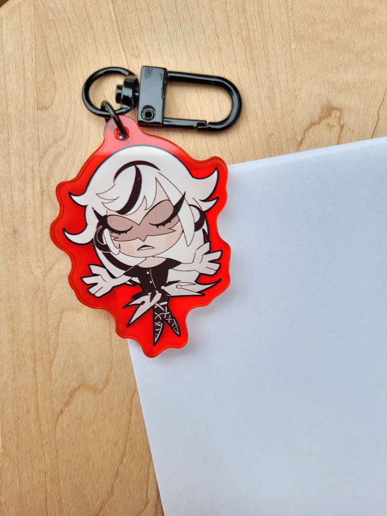 May include: A red acrylic keychain with a cartoon character design. The character has white hair and a black outfit. The keychain has a black clasp and ring. The character has closed eyes and a stern expression. The keychain is on a wooden surface.