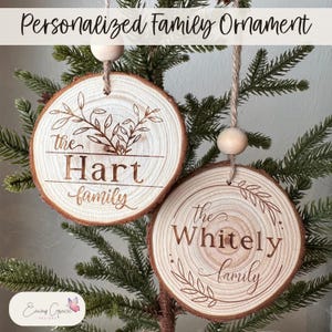 May include: Two wooden ornaments with the text "the Hart family" and "the Whitely family" engraved on them. The ornaments are decorated with a leafy design and are hanging from a tree branch.